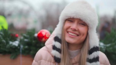 Kış portresi genç yetişkin güzel sarışın kadın komik pofuduk şapkalar giyiyor. Noel havası. Kar yağan kış güzellik moda konsepti. Güzel orta yaşlı kadın Noel çelengi yakınlarında dışarıda oturuyor.