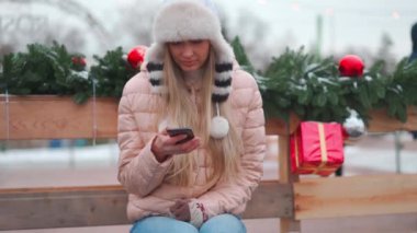 Kış portresi genç yetişkin güzel sarışın kadın komik tüylü şapka takıyor cep telefonu kullanıyor. Noel havası. Kışın kar yağan beyaz kadın elinde akıllı bir telefonla oturmuş gülümsüyor.
