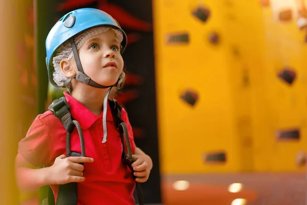 Cute little girl climber in blue protective helmet and gear for climbing standing in climber centre amusement park for children. Caucasian female climber 5 years