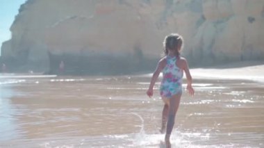 Okyanus sahilinde yaz günü koşan küçük beyaz kız kumsalda koşuyor. Sevimli bayan 5 yaşında. Kumsalda el kamerasından kaçarak eğleniyor. Yüksek kaliteli FullHD görüntüler