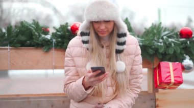 Kış portresi genç yetişkin güzel sarışın kadın komik tüylü şapka takıyor cep telefonu kullanıyor. Noel havası. Kışın kar yağan beyaz kadın elinde akıllı bir telefonla oturmuş gülümsüyor.