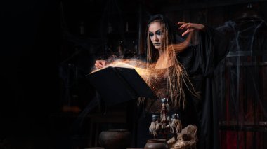 Cadılar Bayramı konsepti Karanlık zindan odasında duran siyah başlıklı cadı büyü yapmak için sihirli kitap kullanıyor. Dişi büyücü büyücü Gotik iç mimari ruh efekti parıldıyor