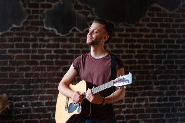 Akustik gitarı olan, tuğla duvarın yanında şarkı söyleyen, hayatın tadını çıkaran yakışıklı, beyaz gitarist ev yapımı müzikal enstrüman çalıyor.