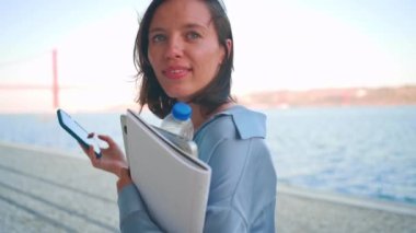 Beyaz tenli güzel bir iş kadını yazın akıllı telefon yürüme seti üzerinden video çekiyor. Avrupalı kadın turist elinde altın rengi dizüstü bilgisayar ve iş kağıdı ile video çekiyor