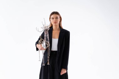 Elinde parlak bir Noel geyiği dekoru tutan genç bir iş kadını beyaz arka planda izole edilmiş siyah takım elbiseli ciddi bir yüzle kameraya bakıyor. Tatil konseptine hazırlanın..