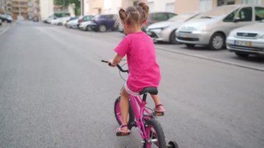 Güneşli bir yaz gününde şehirde bisiklet süren sevimli, gülümseyen bir kız. Çocuklarla aktif aile eğlencesi. Küçük beyaz kız 5 yaşında Pembe çocuk bisikletiyle eğlen