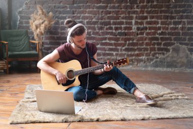 Kafkasyalı genç yetişkin uzak öğretmen bilgisayarlı mobil cihazıyla online gitar çalmayı öğreniyor. Yakışıklı hippi dizüstü bilgisayarla müzik öğretiyor, uzaktan eğitim kavramı 