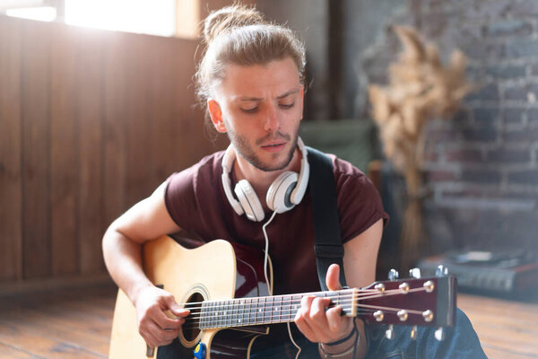 Handsome young man playing acoustic guitar sitting floor living loft room Caucasian male hipster headphones guitar player practicing singing enjoy music relaxing Creative modern young guy Medium shoot