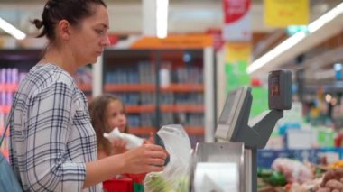 Woman weighs vegetables bought in supermarket on the scales of a self-service store. Mom with child in hypermarket buys vegetables healthy food. Caucasian female use plastic bag for packing 