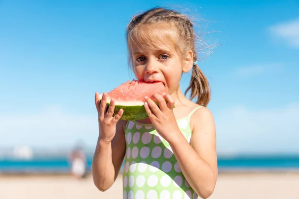 Funny little girl eat watermelon sunny summer day at ocean beach. Cute caucasian female child enjoy summer fruit bite slice of watermelon. Happy childhood. Close up portrait