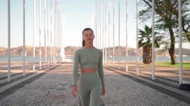 Front view of sportive woman walking on street. Fit caucasian female dressed sportive wear confidence walking on park near seaside sunny summer day. Athletic young woman walk Healthy lifestyle