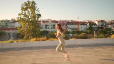 Confident fit female jogging by European city street in beautiful sunny day do fitness training for cardio health. Tracking shot. Slow motion. Handheld. Athletic woman running, morning run