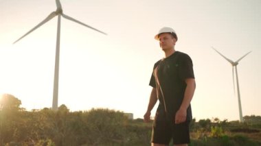Engineer man in white hard hat walking on field with huge windmills during summer sunset. Professional technician. Portrait of an engineer wind turbines. Windmill engineer watching wind turbines 