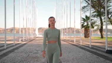 Front view of sportive woman walking on street. Fit caucasian female dressed sportive wear confidence walking on park near seaside sunny summer day. Athletic young woman walk Healthy lifestyle