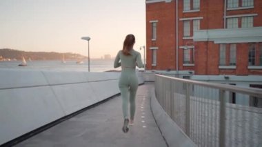 Following Shot of Young Woman in Sportswear Confidently Running through pedestrian Bridge in Summer City Park near Seaside. Back View Athletic Female Jogger run at morning workout training