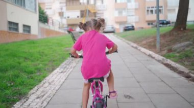 Güneşli bir yaz gününde şehirde bisiklet süren sevimli, gülümseyen bir kız. Çocuklarla aktif aile eğlencesi. Küçük beyaz kız 5 yaşında Pembe çocuk bisikletiyle eğlen
