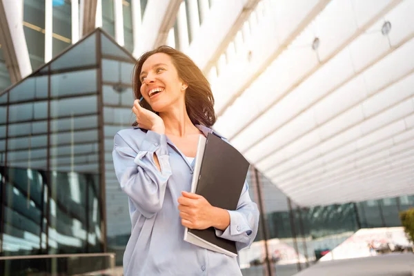 Attractive brunette businesswoman talking mobile phone with client. Pretty caucasian female business person smile when phone conversation. Woman managing business talking via mobile phone standing