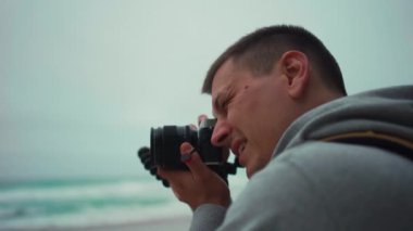 Hipster traveler photographer takes a photo beautiful Atlantic Ocean landscape on professional camera on evening dusk time. Man tourist with yellow backpack shoots photo seascape. Handheld effect