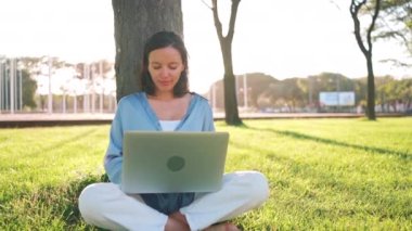 Parktaki çimlerin üzerinde otururken dizüstü bilgisayar kullanan mutlu esmer kadın. Parkta lotus pozisyonunda oturan genç Avrupalı kadın dizüstü bilgisayar kullanıyor. 