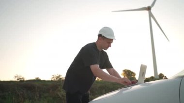 Technician engineer working at wind turbine using laptop computer worker or operator checks wind turbine farm does maintenance to power generator station lean on car wind turbine and sunset background