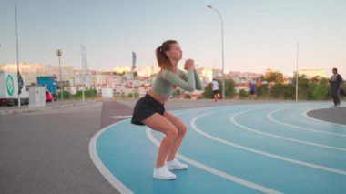 Koşu pistindeki sportif kadın çömelme egzersizleri yapıyor. Vücut ağırlığı açık hava eğitimi. Genç yetişkin bir kadın dışarıda egzersiz yapıyor. Avrupalı kadın çömeliyor. Atletik Kafkas kadın sporu.