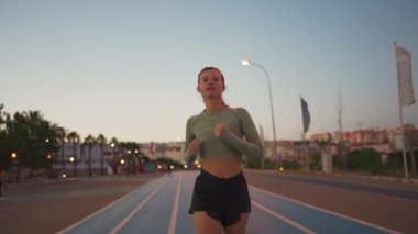 Spor sahasının yakınındaki pistte koşan bayan sporcu. Akşamüstü koşu pistinde koşan, zinde, beyaz bir kadın. Atletik bayan koşucu dışarıda antrenman yapıyor. Sağlıklı yaşam tarzı kavramı