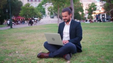 Handsome hispanic male businessman sitting lotus position in public park use laptop. Remote work concept. Stylish freelancer in suit relax in park summer day working on computer. Technology. Handheld.