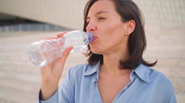 Kendine güvenen bir kadın yaz günü ayakta duran plastik şişe su içer. Beyaz, orta yaşlı, mavi gömlekli bayan iş adamı. Suyu dışarı çıkarıyor. Başka tarafa bakıyor ve gülümsüyor.. 
