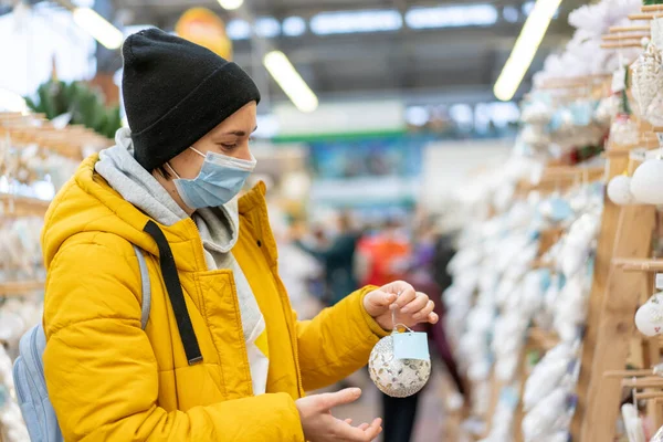 Christmas decoration shopping. Caucasian woman choose new year decor in shopping mall. Medium age female dressed medical mask and yellow winter parka buying Christmas ornament toys indoor store
