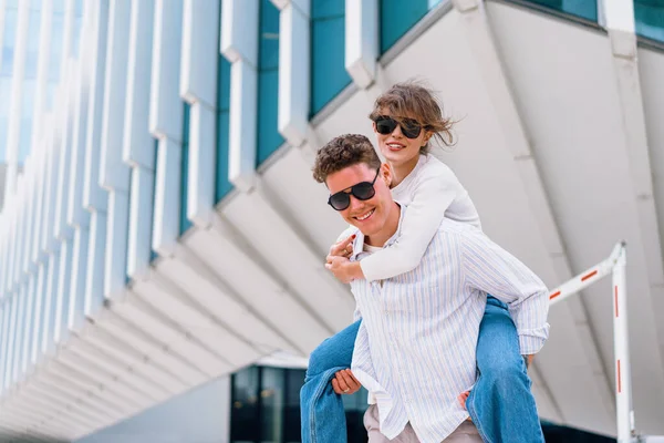带着太阳镜的无忧无虑的城市年轻夫妇们在城市的大街上背负着现代商业建筑 男人背着年轻女人 积极向上的情绪在散步时很开心 — 图库照片