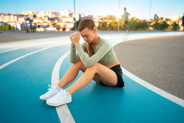 Tired upset female athlete sitting on running track resting after jogging exercises. Disappointed sport result. Amateur sprinter sportswoman exhausted after tough trainings sitting on track