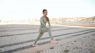 Fitness woman 20s in sports wear doing lunges on step, and stretching legs on summer park near seaside. Warm up before jogging workout. Athletic female jogger training legs. Squat lunges training