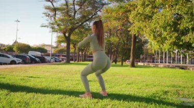 Sporty woman in park squats. Bodyweight training open air. Young adult fitness woman exercising outdoor on green grass. European woman squats. Athletic caucasian female person workout outside
