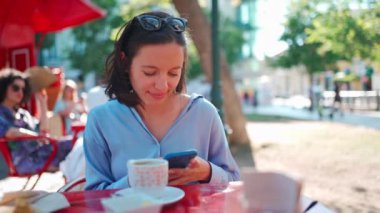 Pretty woman sitting outdoors cafeteria use smartphone chatting with friends. Beautiful caucasian business woman coffee break. Front view portrait shoot. Girl with sunglasses on head resting cafe 