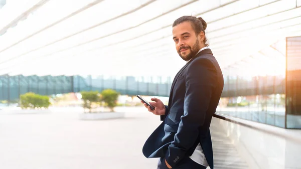 Businessman standing outdoor use smartphone looking camera screen confident. Handsome hispanic male business person in business suit holding smartphone in hand. Modern business person low angle view