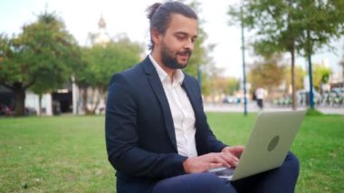 Handsome hispanic male businessman sitting lotus position in public park use laptop. Remote work concept. Stylish freelancer in suit relax in park summer day working on computer. Technology. Handheld.