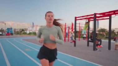 Spor sahasının yakınındaki pistte koşan bayan sporcu. Akşamüstü koşu pistinde koşan, zinde, beyaz bir kadın. Atletik bayan koşucu dışarıda antrenman yapıyor. Sağlıklı yaşam tarzı kavramı