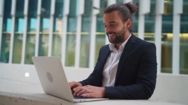 Hispanic businessman standing outside with laptop working remote distant job outdoors near office company building. Stylish unshaved male business man with long hair dressed business suit use laptop