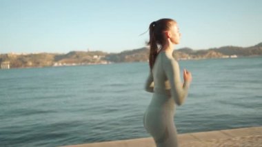 Confident fit female jogging by embankment area coastal city in beautiful sunny day do fitness training for cardio health. Tracking shot. Slow motion. Handheld. Athletic woman running, morning run