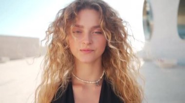 Açık havada kıvırcık kadın portresi. Sarışın, kıvırcık bir kadının kameraya ciddi ciddi bakıp gülümsediği yakın çekim. Mutlu olumlu duygular. Genç kadın, beyaz tenli. Saygıdeğer yüz ifadesi 