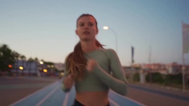 Spor sahasının yakınındaki pistte koşan bayan sporcu. Akşamüstü koşu pistinde koşan, zinde, beyaz bir kadın. Atletik bayan koşucu dışarıda antrenman yapıyor. Sağlıklı yaşam tarzı kavramı