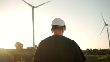 Engineer man in white hard hat walking on field with huge windmills during summer sunset. Professional technician. Portrait of an engineer wind turbines. Windmill engineer watching wind turbines 