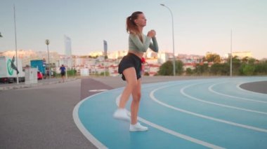 Koşu pistindeki sportif kadın çömelme egzersizleri yapıyor. Vücut ağırlığı açık hava eğitimi. Genç yetişkin bir kadın dışarıda egzersiz yapıyor. Avrupalı kadın çömeliyor. Atletik Kafkas kadın sporu.