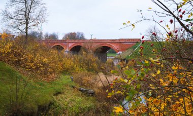 Nehir üzerinde eski bir kırmızı tuğla köprü, ön planda sonbahar manzarası, sarı yapraklardan çalılar..