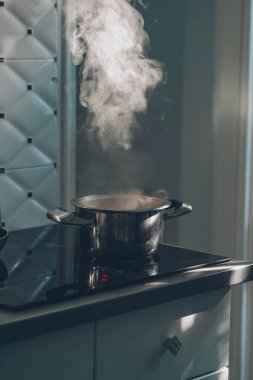The process of cooking soup in a metallic pot on an electric stove. Evening light in the kitchen. Beautiful hot steam is rising from a saucepan. Sun rays from window in the cuisine. Universal colors