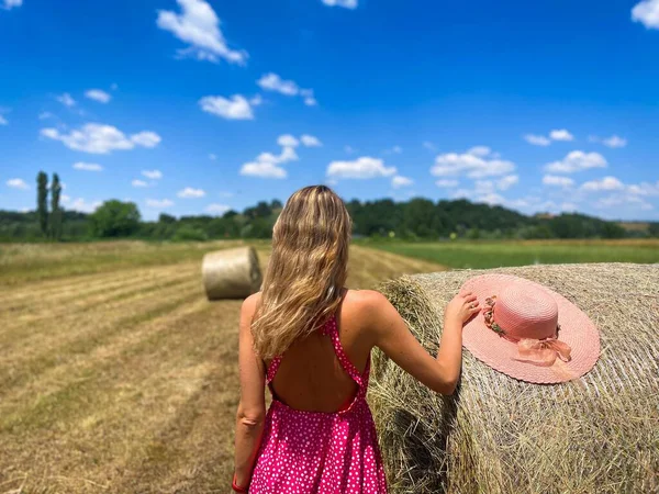 Woman with pink summer dress and hat standing near bales of hay in the middle of a field during a hot summer day