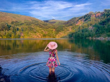 Güzel bir manzarayla çevrili, şapkalı ve elbiseli bir kadın göle giriyor.