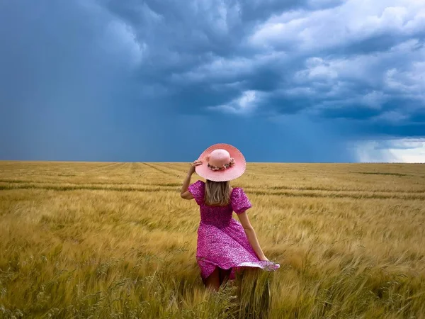 Woman in pink dress and hat in a field of wheat on a cloudy and stormy summer day