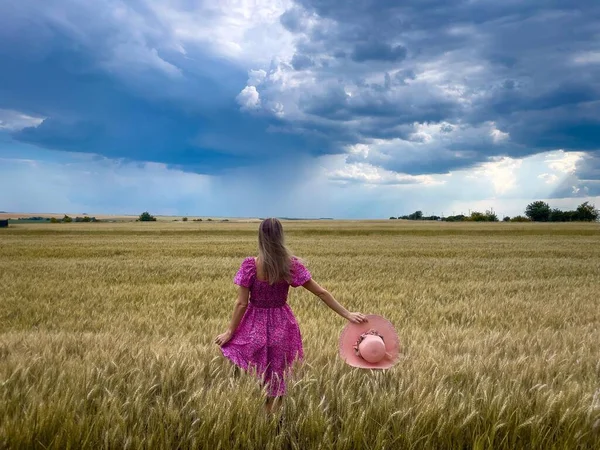 Woman in pink dress and hat in a field of wheat on a cloudy and stormy summer day