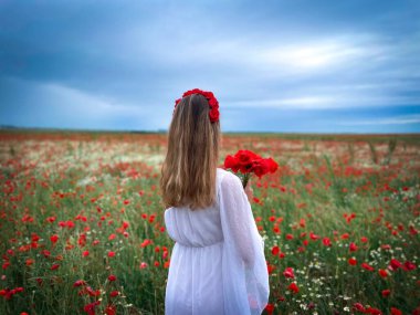 Young blonde woman in white dress standing in a field of poppies on a cloudy summer day
